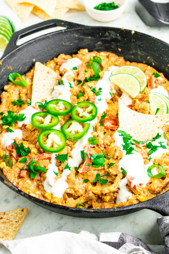 Overhead view of a cast iron skillet filled with creamy silken tofu sour cream corn and soy curls topped with sliced jalapenos and lime wedges surrounded by tortilla chips