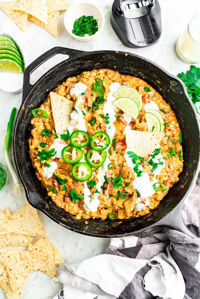 Overhead view of a cast iron skillet filled with creamy silken tofu sour cream corn and soy curls topped with sliced jalapenos and lime wedges surrounded by tortilla chips