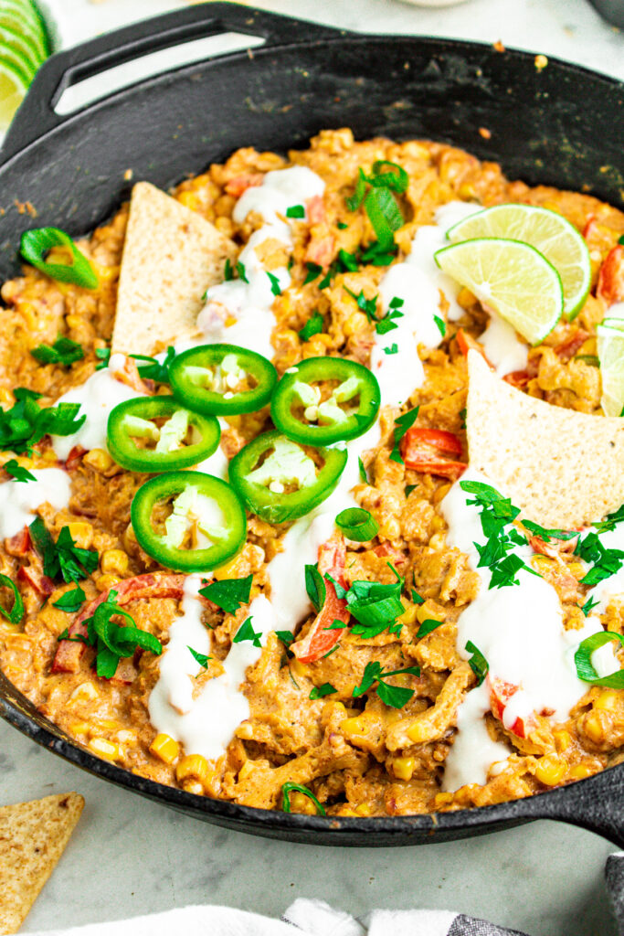 Overhead close up view of a cast iron skillet filled with creamy vegan sour cream corn and soy curls topped with sliced jalapenos and lime wedges surrounded by tortilla chips