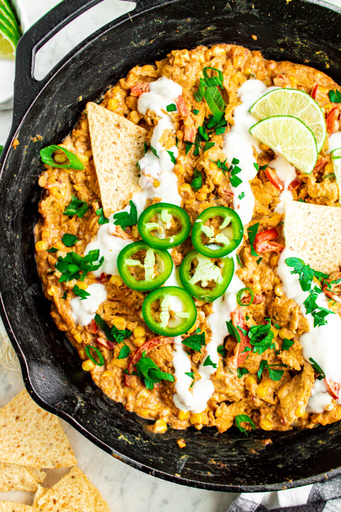 Overhead view of a cast iron skillet filled with creamy vegan sour cream corn and soy curls topped with sliced jalapenos and lime wedges surrounded by tortilla chips