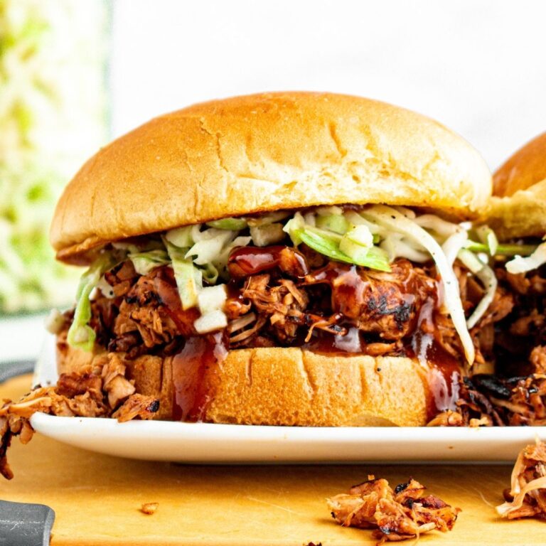 Close up head on view of a delightfully messy BBQ vegan pulled pork sandwich stuffed with jackfruit pulled pork on a toasted bun with green cabbage slaw and extra BBQ sauce dripping down the sides