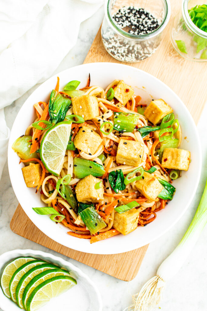Overhead view of a round white bowl filled with ginger sesame noodles and crispy tofu cubes next to a dish of lime wedges and a jar of black and white sesame seeds