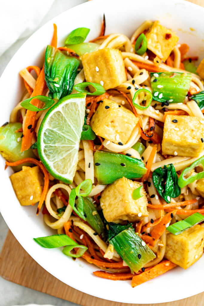Close up overhead view of a round white bowl filled with garlic ginger sesame noodles with crispy tofu cubes, bok choy, carrot strips, and lime wedges