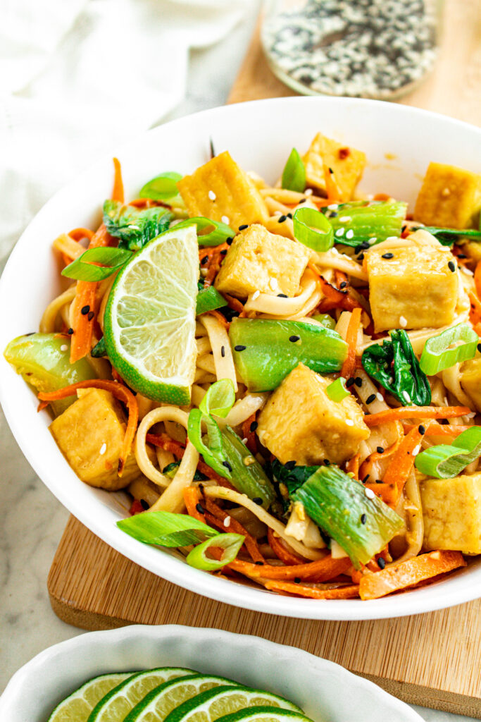 Overhead view of a round white bowl filled with ginger sesame noodles and crispy tofu cubes next to a dish of lime wedges and a jar of black and white sesame seeds