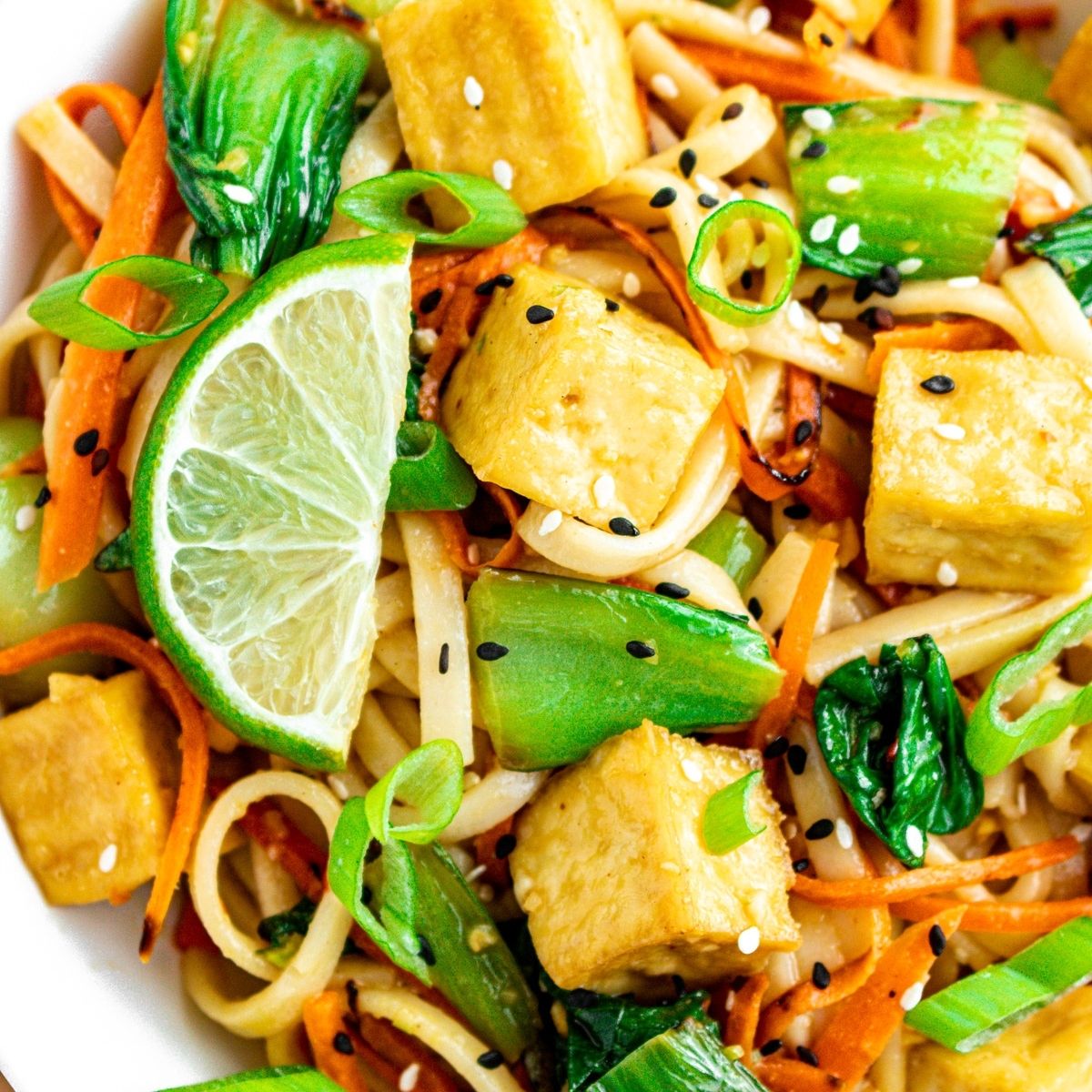 Close up overhead view of a round white bowl filled with garlic ginger sesame noodles with crispy tofu cubes, bok choy, carrot strips, and lime wedges