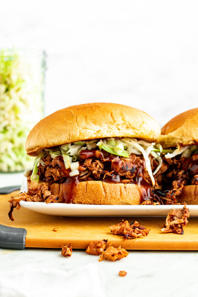 Head on view of a messy vegan pulled pork sandwich stuffed with jackfruit pulled pork on a toasted bun with green cabbage slaw