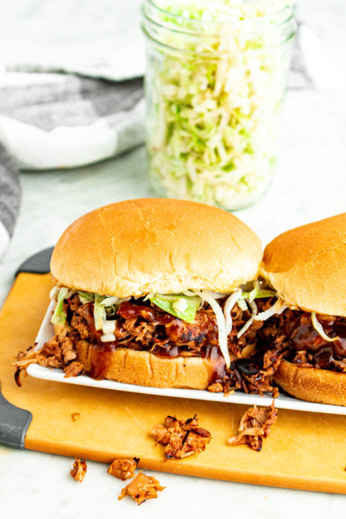 Overhead view of two beautifully messy vegan pulled pork sandwiches stuffed with jackfruit pulled pork on a toasted bun with green cabbage slaw and BBQ sauce drizzling down the sides