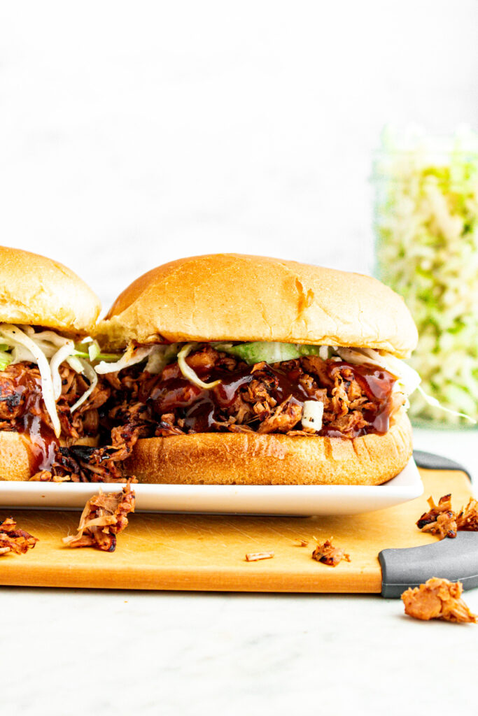 Head on view of a magnificently messy vegan pulled pork sandwich stuffed with jackfruit pulled pork on a toasted bun with green cabbage slaw and BBQ sauce drizzling down the sides