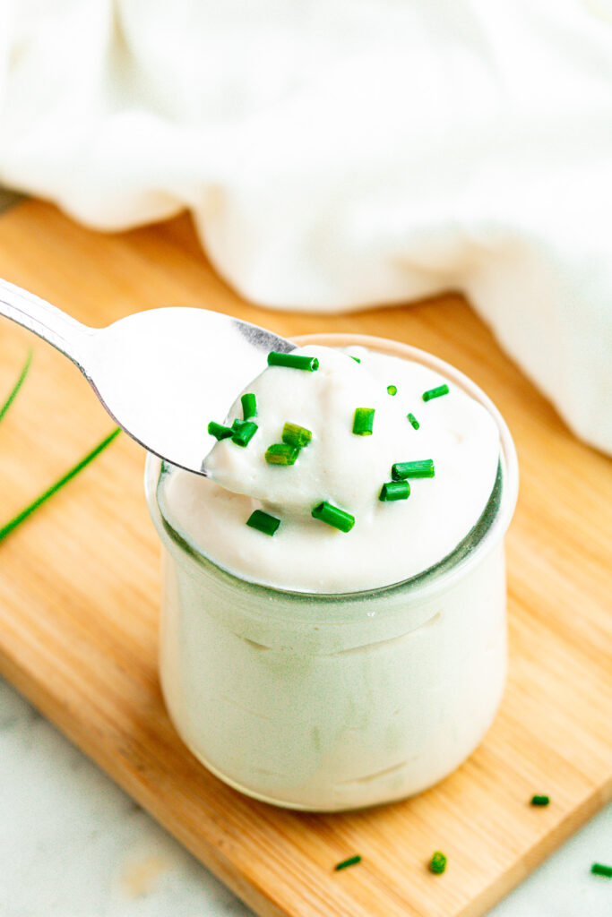 Overhead view of a clear glass jar filled with creamy vegan sour cream topped with chives with a silver spoon digging into it