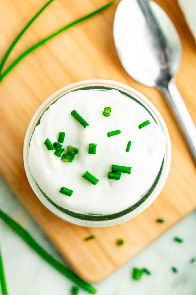 Overhead view of a clear glass jar of creamy vegan sour cream topped with chopped green onions on a wood cutting board next to a silver spoon
