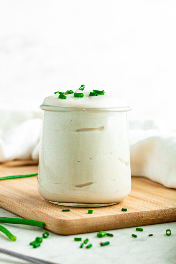 Head on view of a clear glass jar overflowing with vegan sour cream and topped with chopped green onions on a wood cutting board