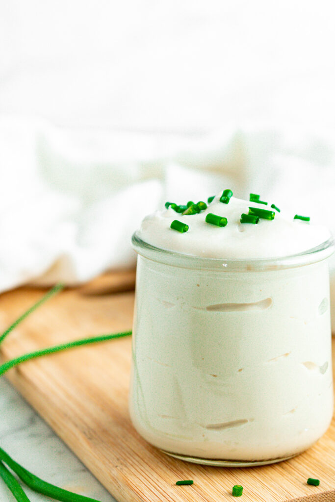 Head on view of a clear glass jar overflowing with vegan sour cream and topped with chopped green onions on a wood cutting board