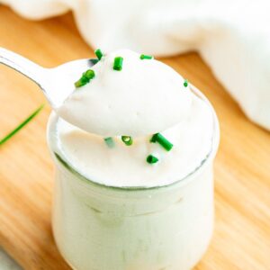 Overhead view of a clear glass jar filled with creamy vegan sour cream topped with chives with a silver spoon digging into it