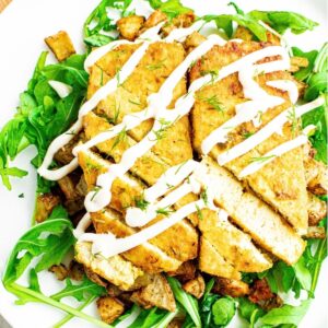 Overhead view of two sliced dill pickle tofu cutlets on a bed of crispy potatoes and arugula drizzled with vegan sour cream