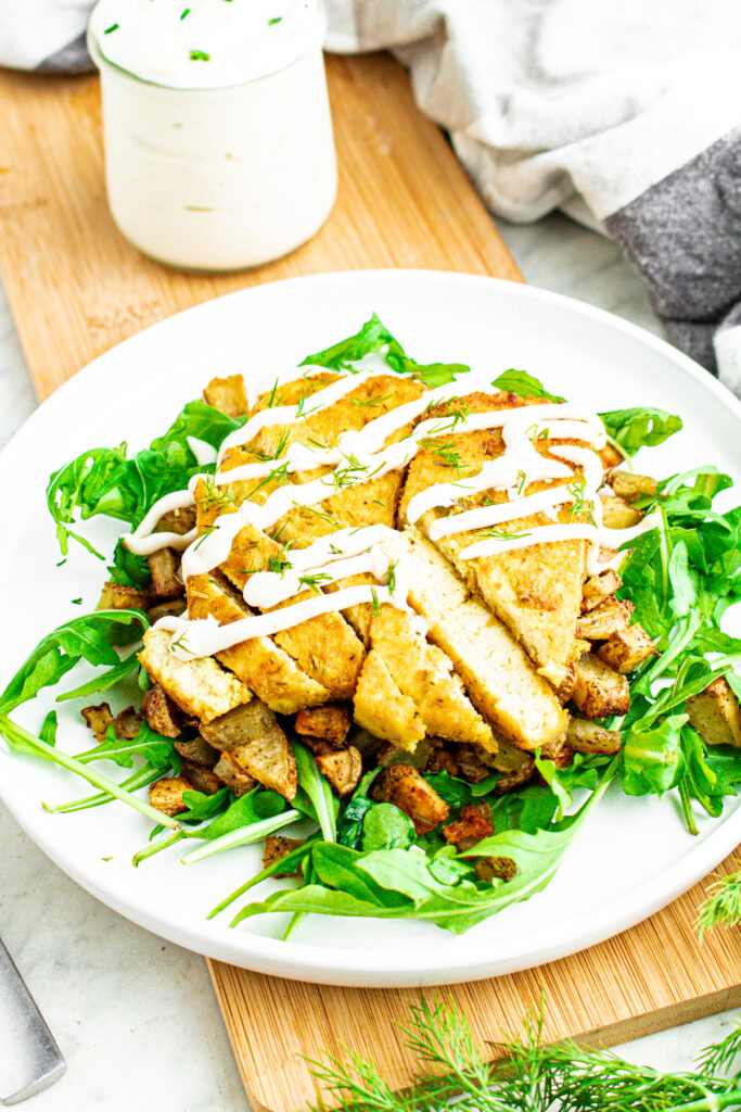 Overhead view of two sliced dill pickle tofu cutlets on a bed of crispy potatoes and arugula drizzled with vegan sour cream