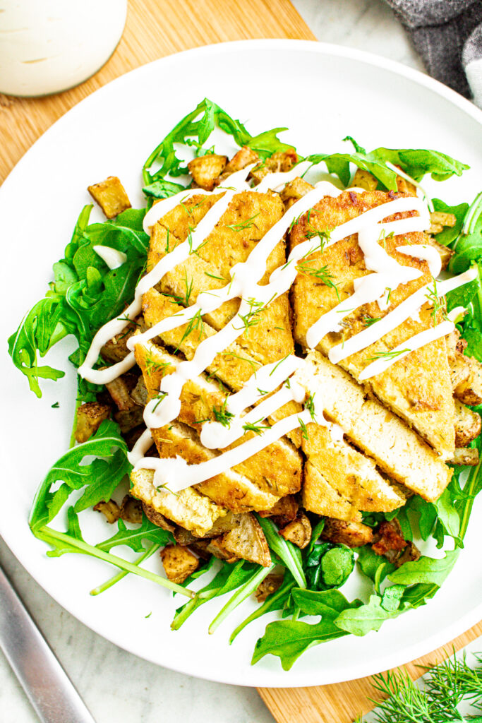 Overhead view of two sliced dill pickle tofu cutlets on a bed of crispy potatoes and arugula drizzled with vegan sour cream