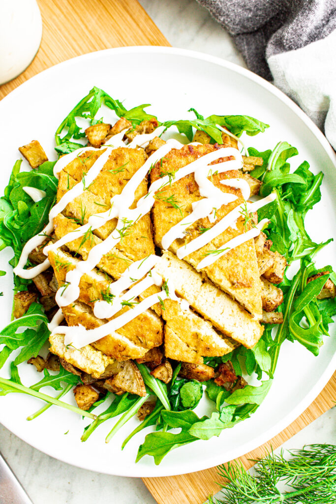 Overhead view of two sliced dill pickle tofu cutlets on a bed of crispy potatoes and arugula drizzled with vegan sour cream